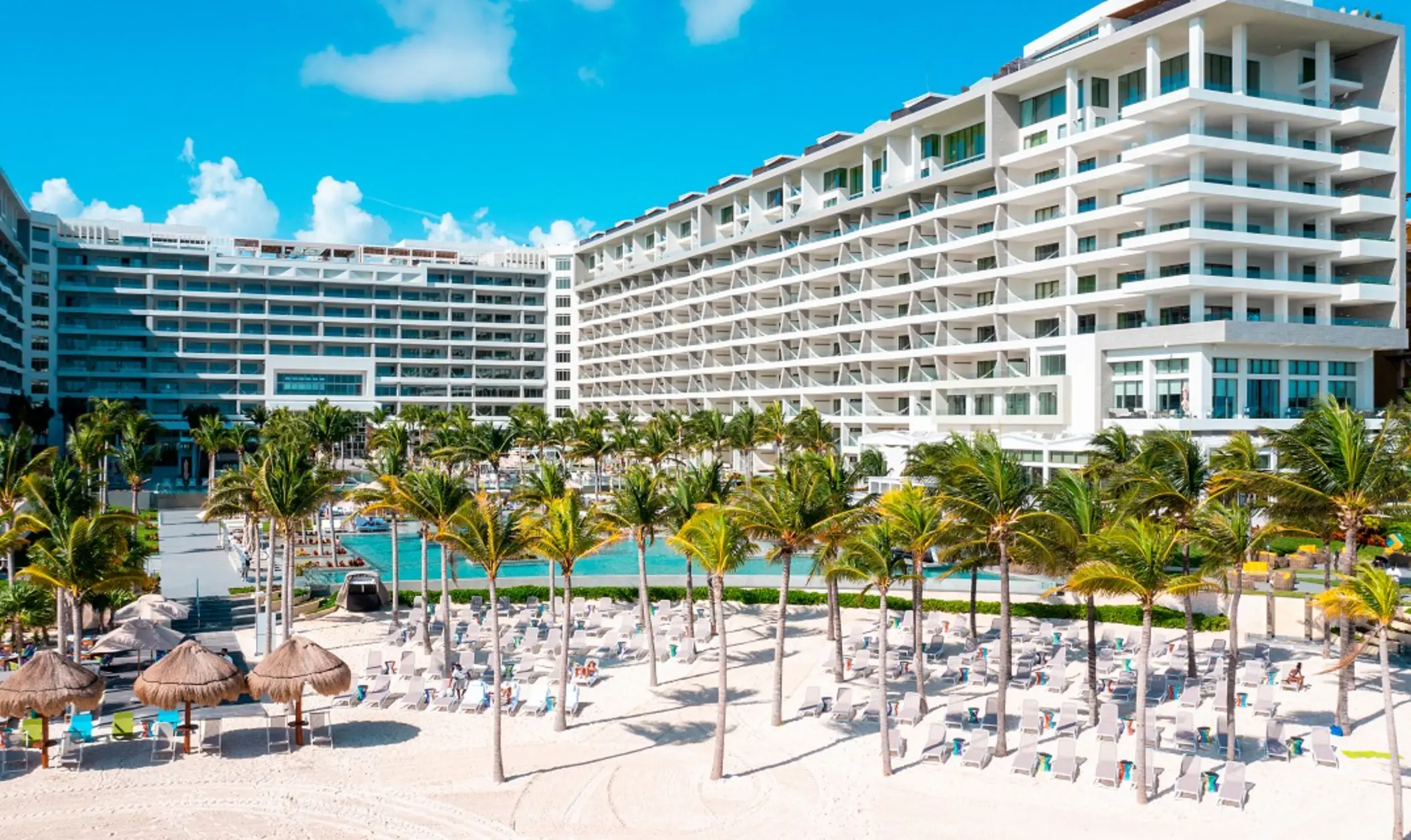 Beachfront resort with a wide modern facade, lined with palm trees, sun loungers, and oceanfront pools.