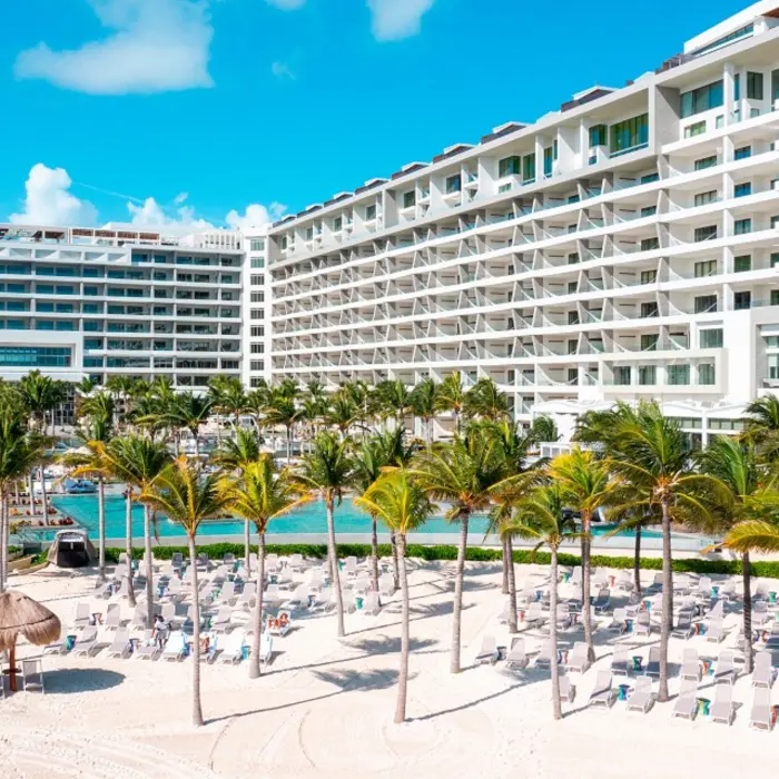 Beachfront resort with a wide modern facade, lined with palm trees, sun loungers, and oceanfront pools.