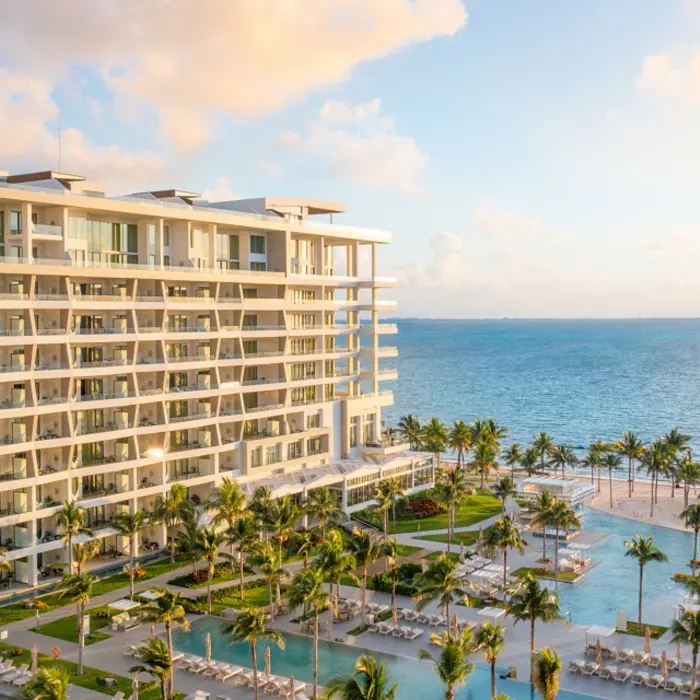 Resort view at sunset with ocean backdrop, palm trees, and multiple pools surrounded by loungers and greenery.