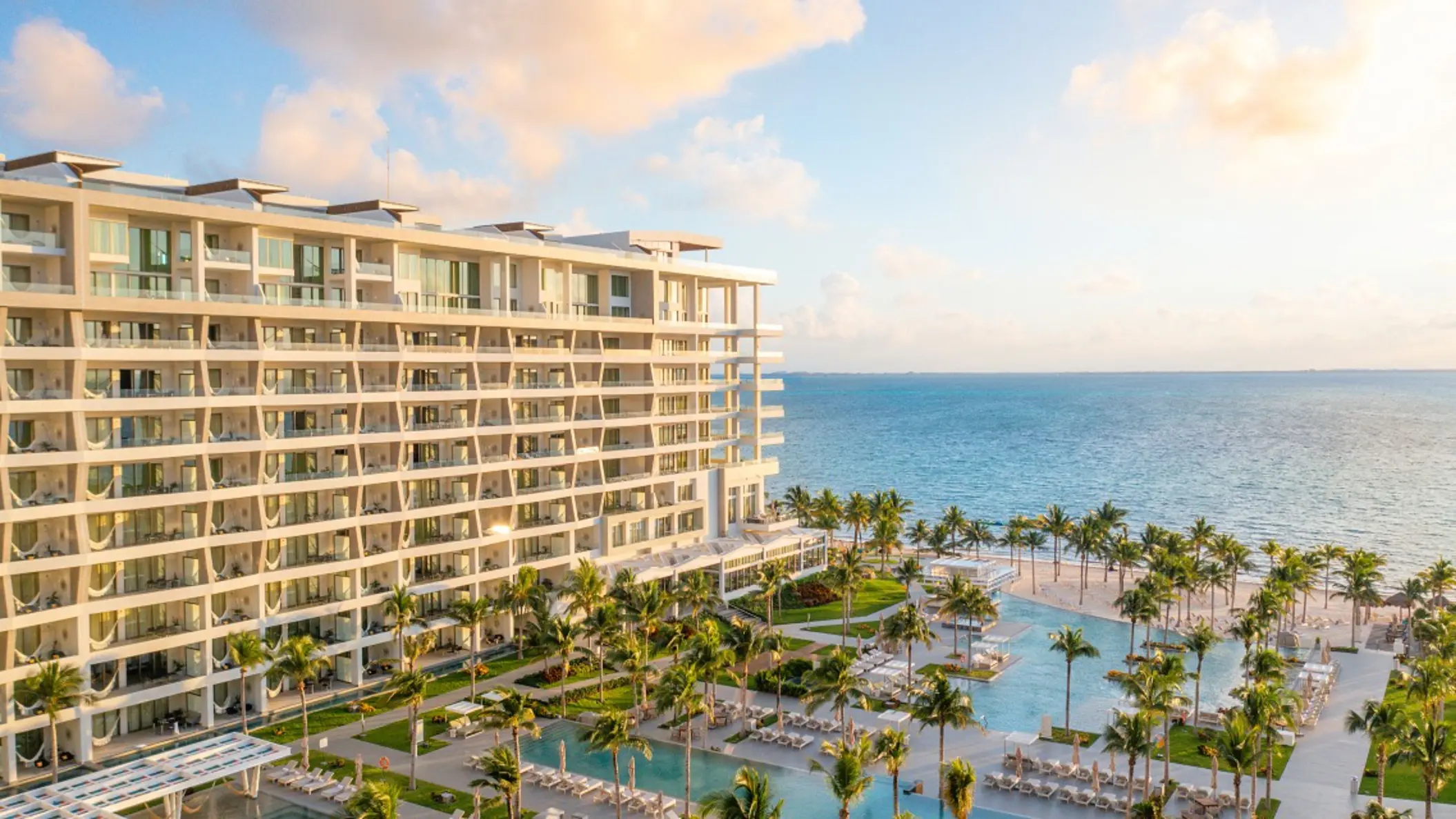 Resort view at sunset with ocean backdrop, palm trees, and multiple pools surrounded by loungers and greenery.