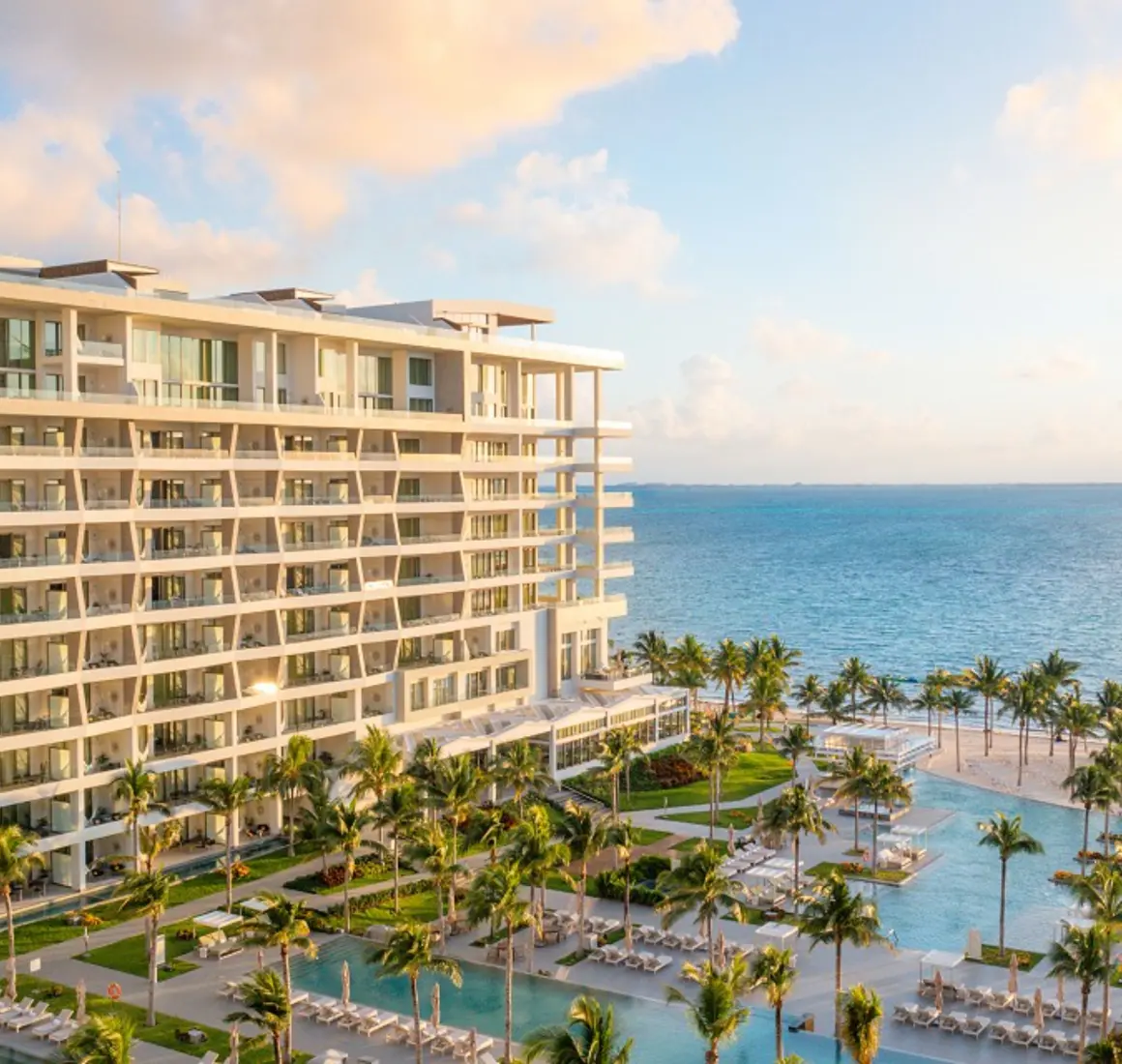 Resort view at sunset with ocean backdrop, palm trees, and multiple pools surrounded by loungers and greenery.