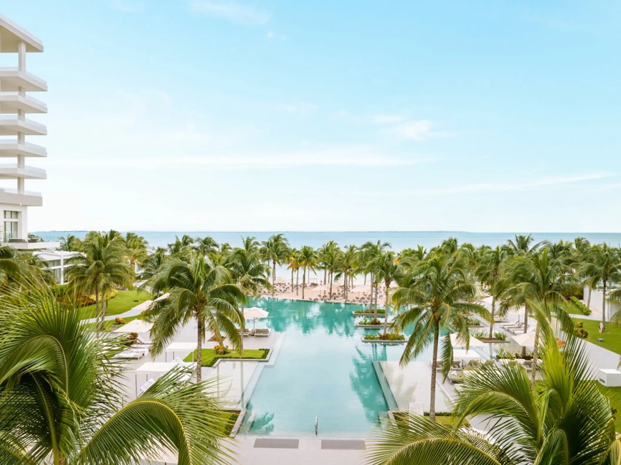 Tropical aerial view featuring palm-lined pools, white beach loungers, and ocean waters under blue skies.