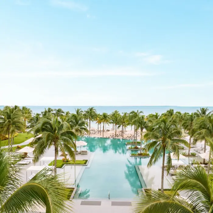 Tropical aerial view featuring palm-lined pools, white beach loungers, and ocean waters under blue skies.