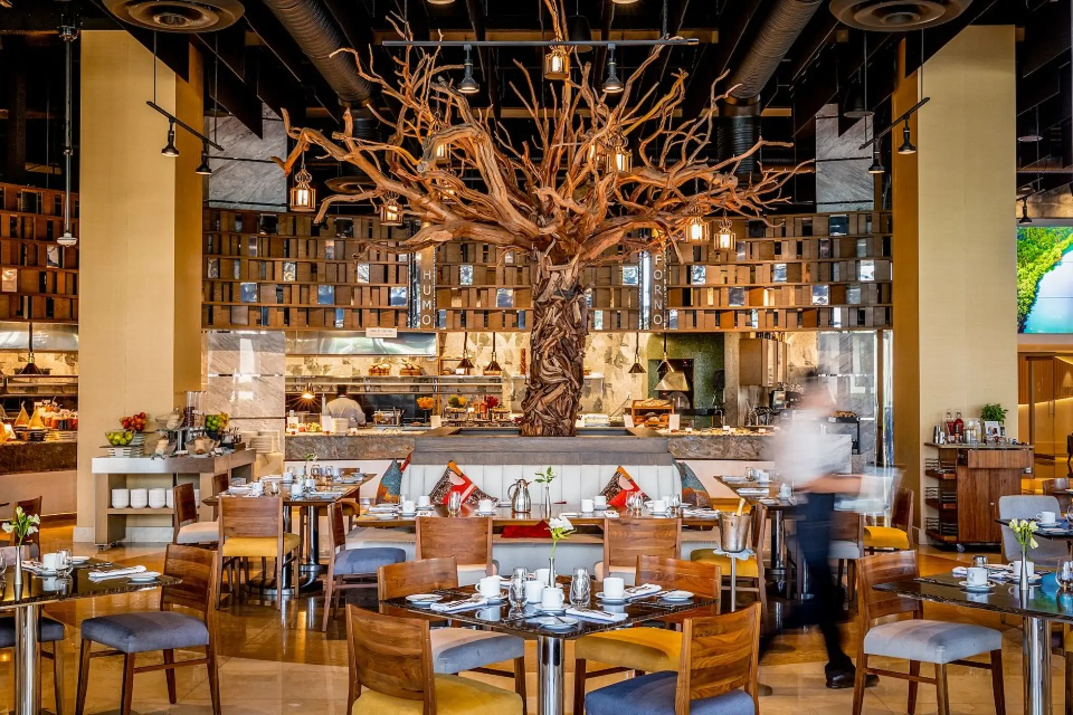 Upscale restaurant with a dramatic sculpted tree centerpiece, open kitchen, and cozy wood-accented dining setup.