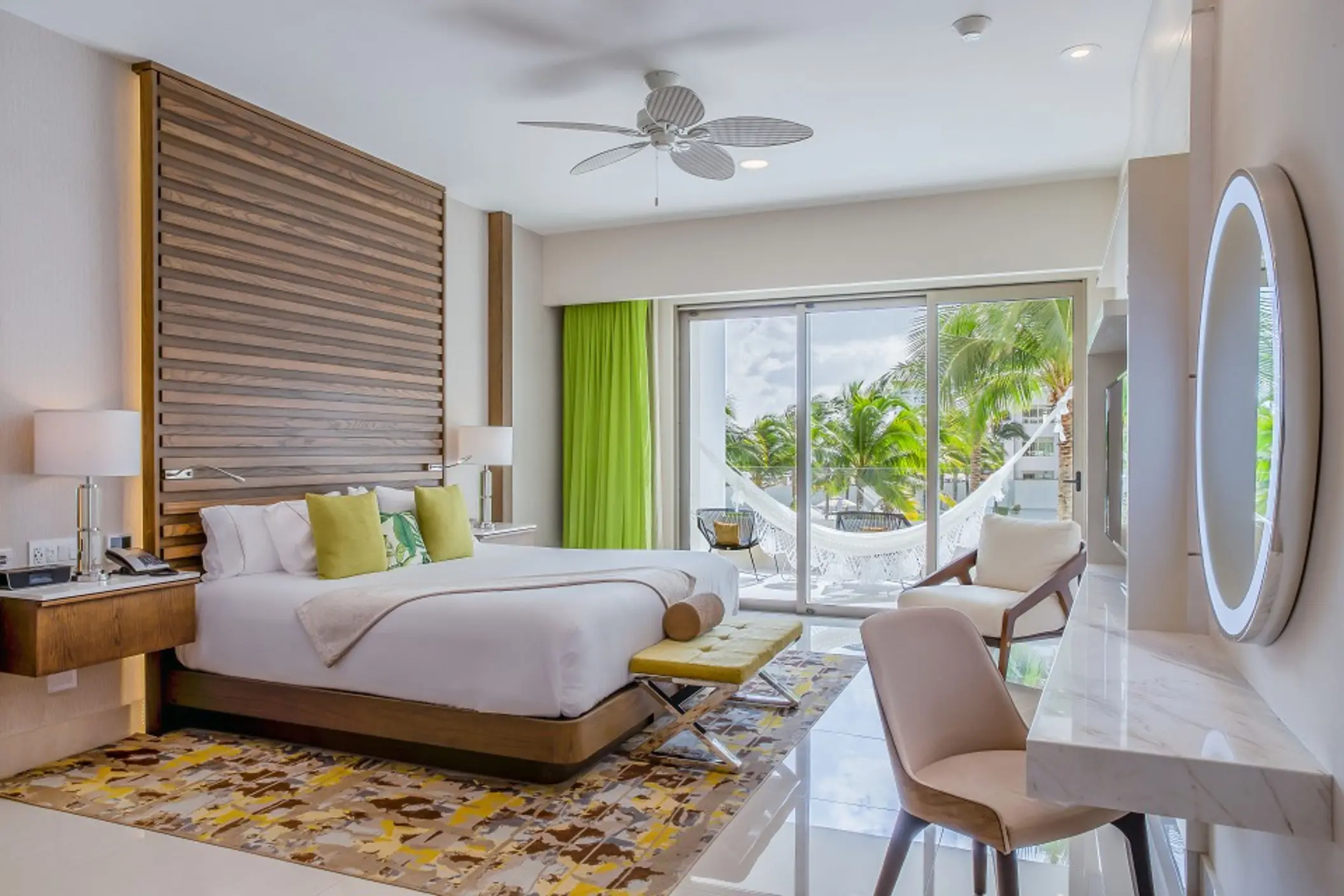 Elegant bedroom with king bed, tropical pillows, hammock on balcony, and view of lush palm trees outside.