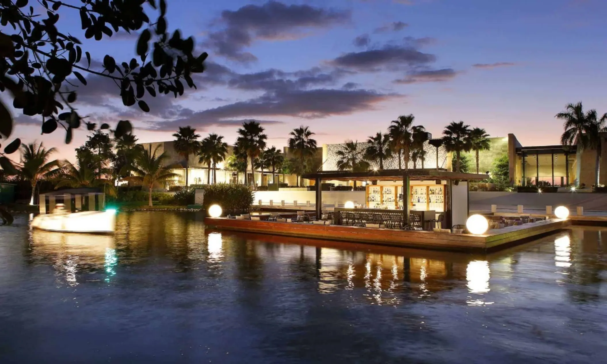 Floating outdoor bar surrounded by water and palm trees, glowing warmly at dusk in a tropical setting.