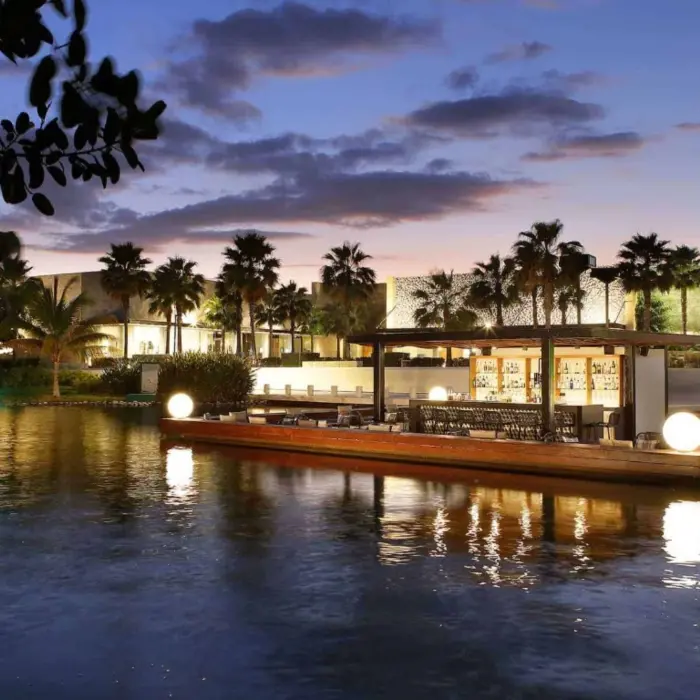 Floating outdoor bar surrounded by water and palm trees, glowing warmly at dusk in a tropical setting.