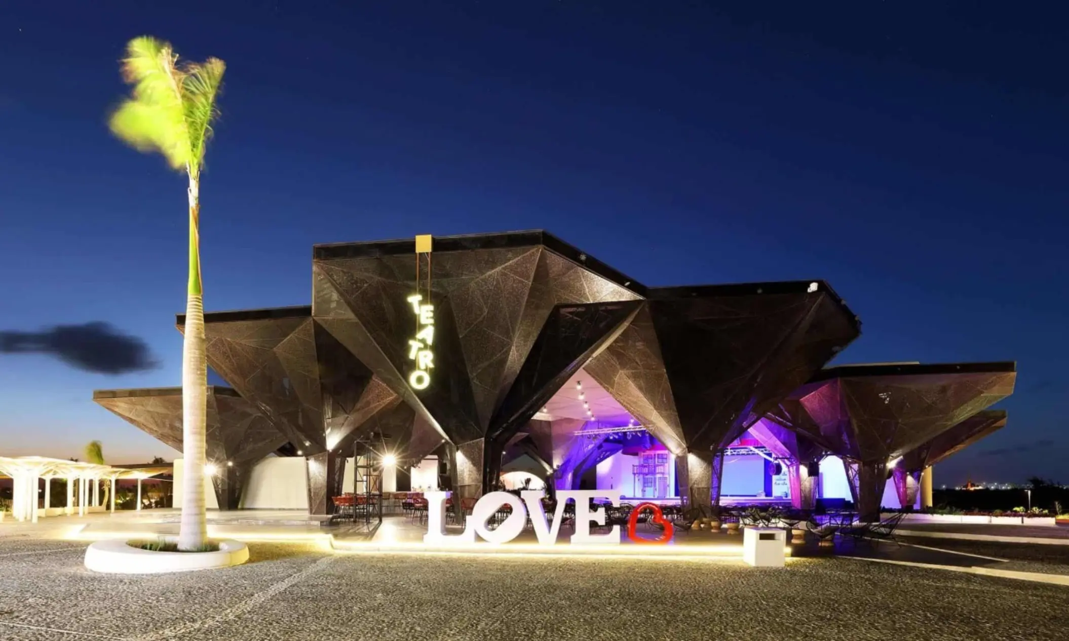 Contemporary theater building with glowing 'Teatro' sign and LOVE sculpture at night.