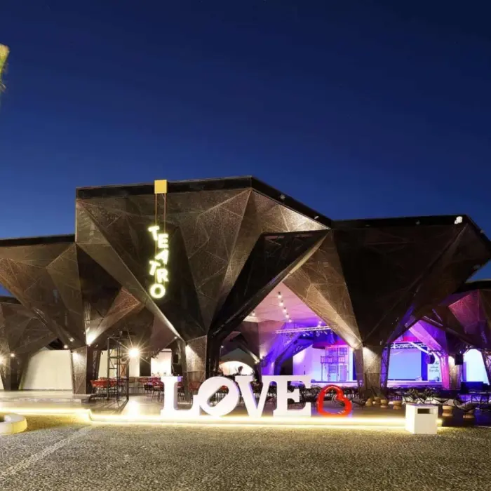 Contemporary theater building with glowing 'Teatro' sign and LOVE sculpture at night.