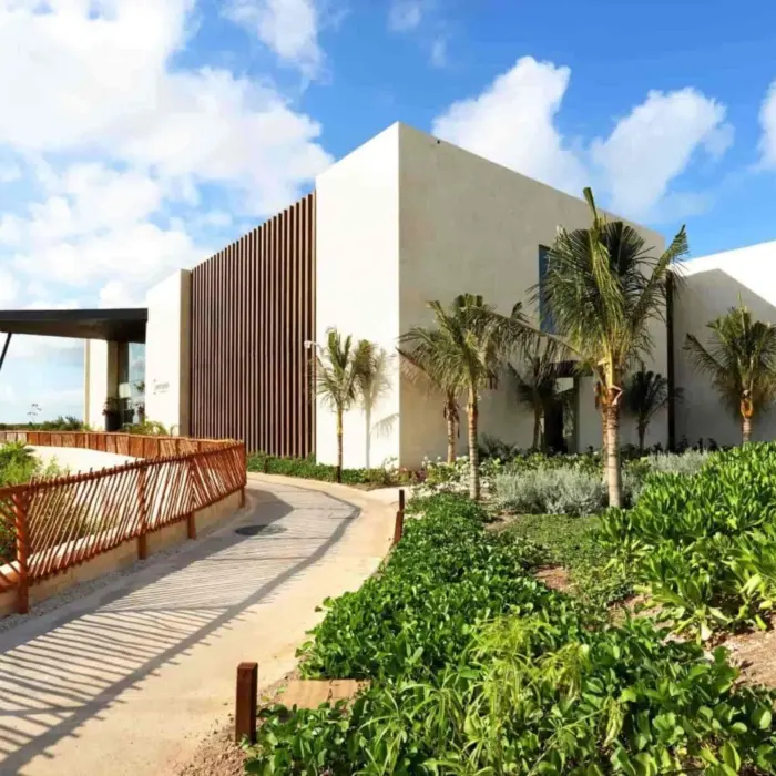 Modern building entrance surrounded by lush greenery and palm trees under a bright blue sky.