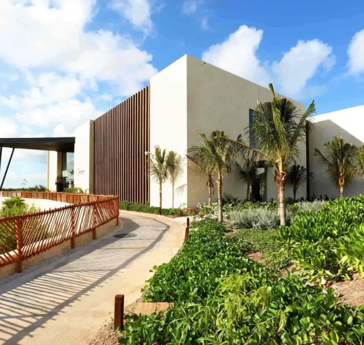 Modern building entrance surrounded by lush greenery and palm trees under a bright blue sky.