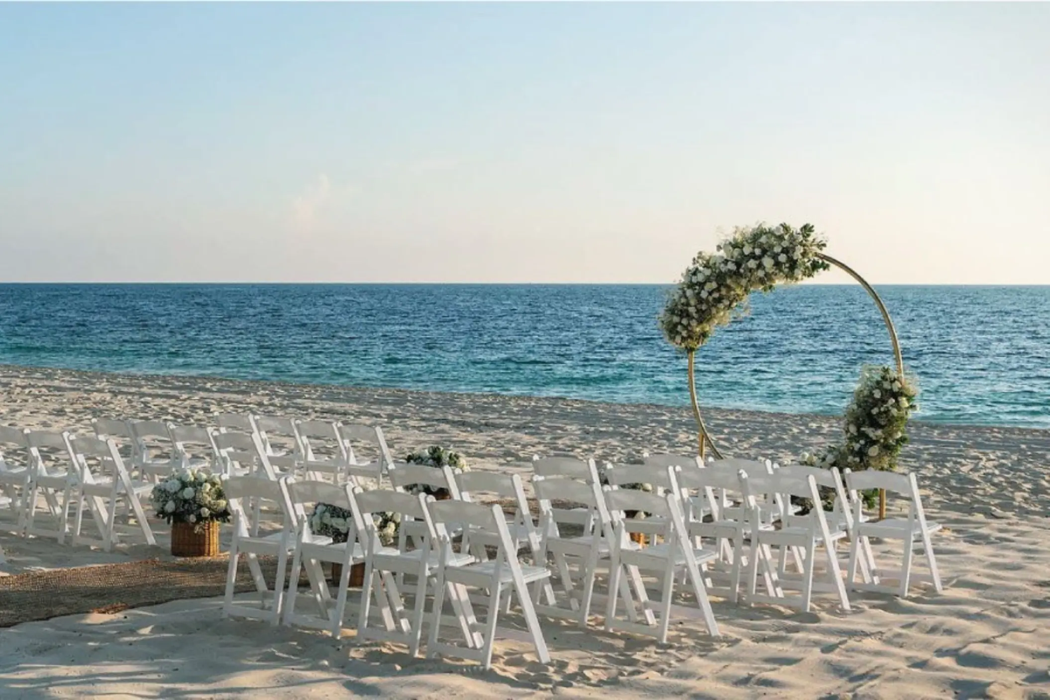 Beach wedding setup with white chairs and a circular floral arch facing the calm, turquoise ocean.