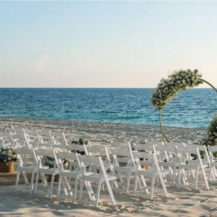 Beach wedding setup with white chairs and a circular floral arch facing the calm, turquoise ocean.