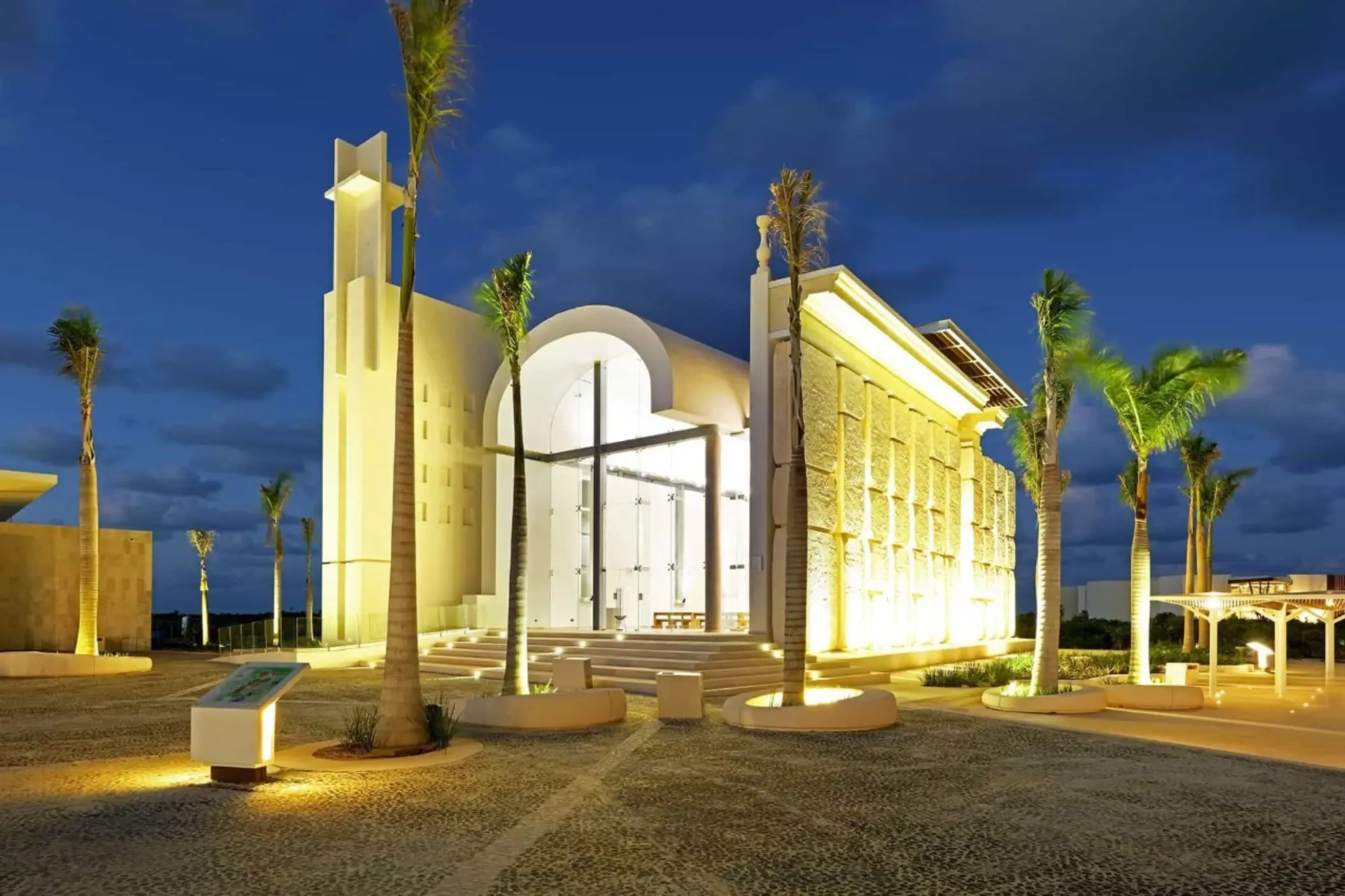 Grand illuminated chapel with tall palm trees and modern architectural design at twilight.