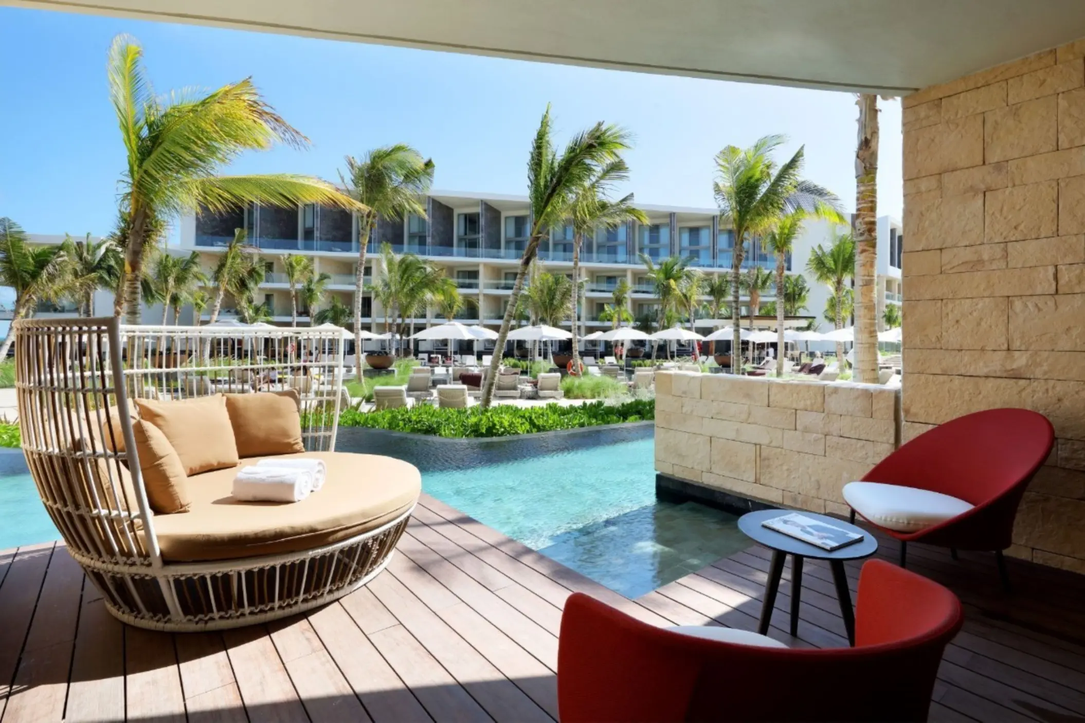 Swim-out suite patio with daybed and red chairs, overlooking tropical pool area and palm trees.