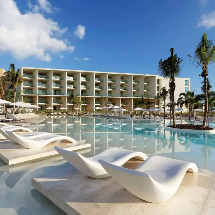 Resort pool with curved white loungers and lush landscaping in front of modern beachfront buildings.