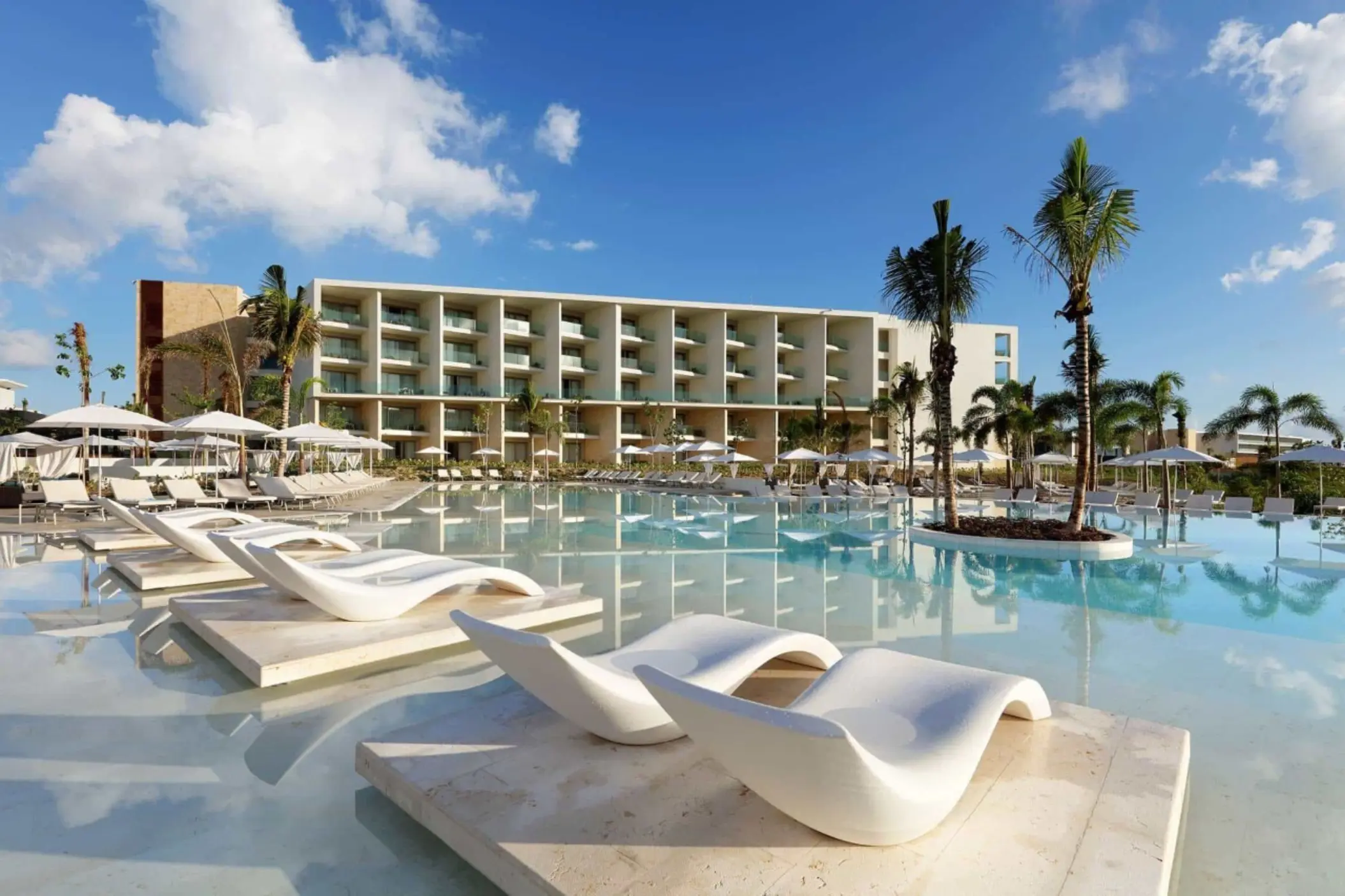Resort pool with curved white loungers and lush landscaping in front of modern beachfront buildings.