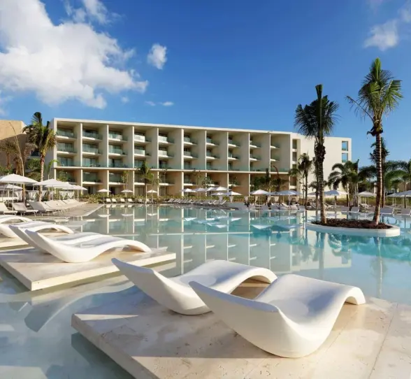 Resort pool with curved white loungers and lush landscaping in front of modern beachfront buildings.
