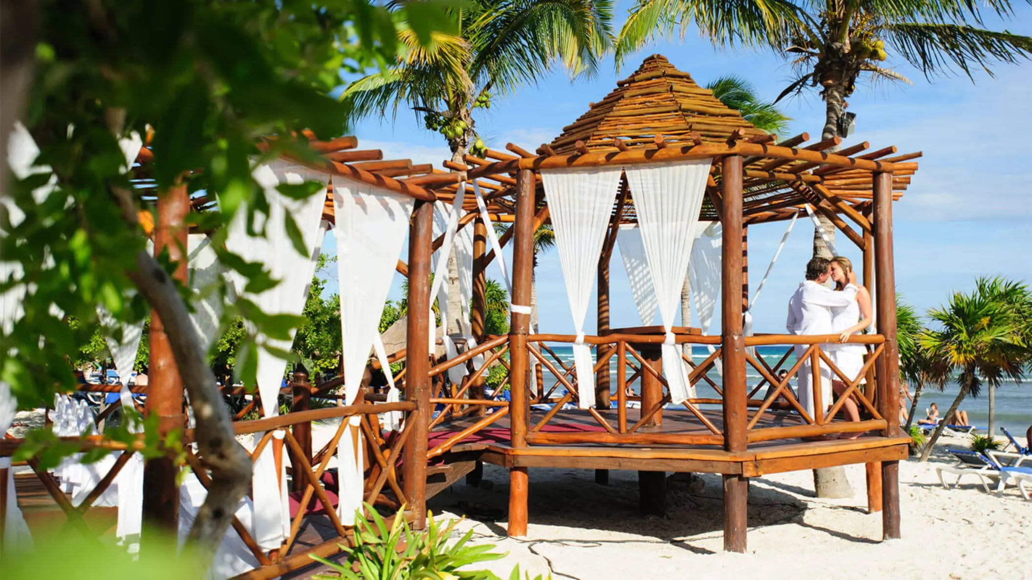 Bride and groom embracing in a picturesque wooden gazebo venue on a sandy beach, surrounded by palm trees.