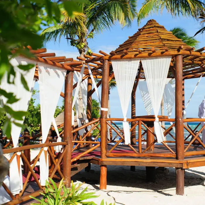 Bride and groom embracing in a picturesque wooden gazebo venue on a sandy beach, surrounded by palm trees.