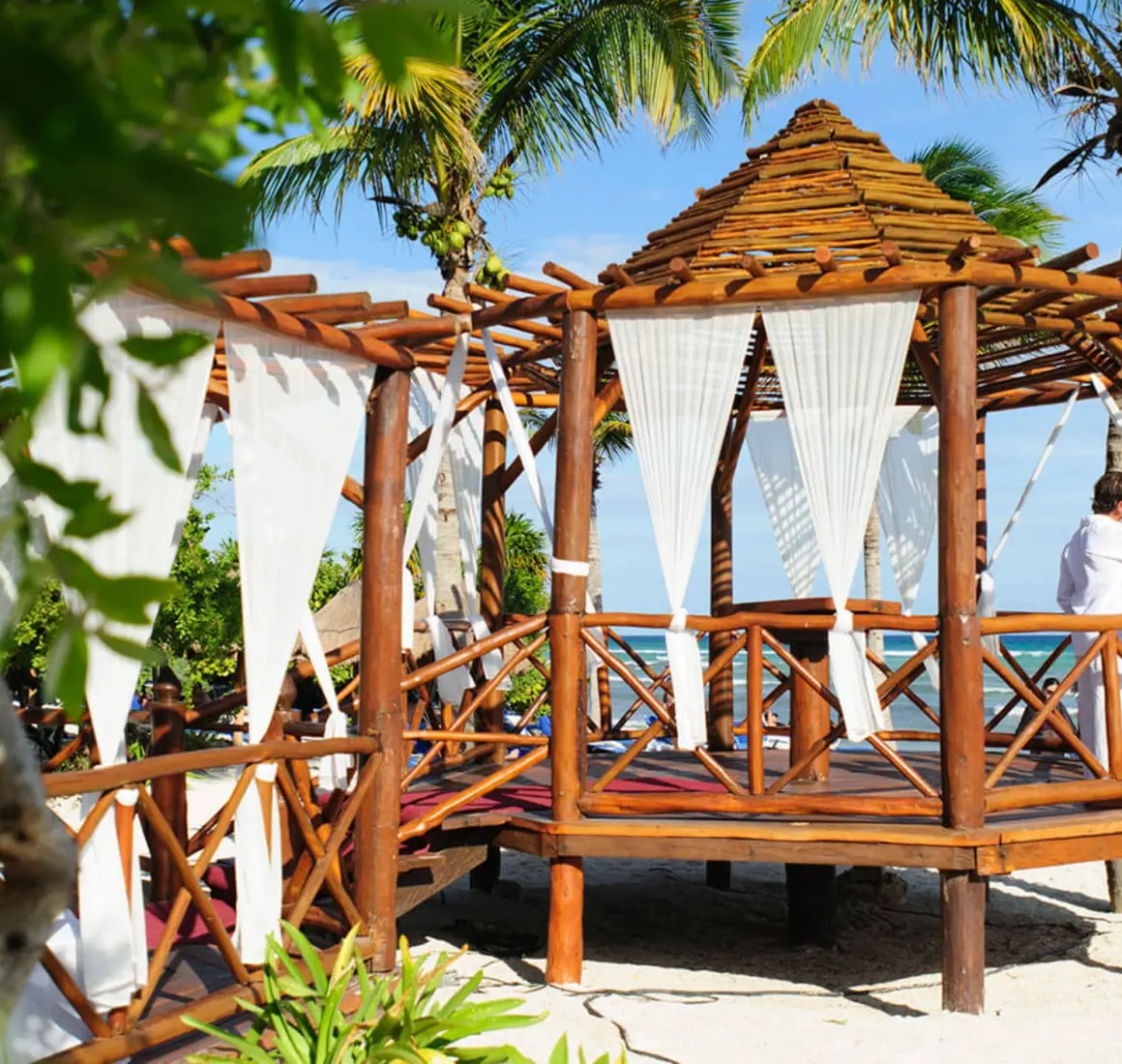 Bride and groom embracing in a picturesque wooden gazebo venue on a sandy beach, surrounded by palm trees.