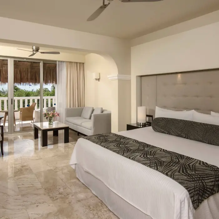 Spacious Platinum Suite at Grand Riviera Princess with a large bed, seating area, and balcony overlooking tropical foliage.