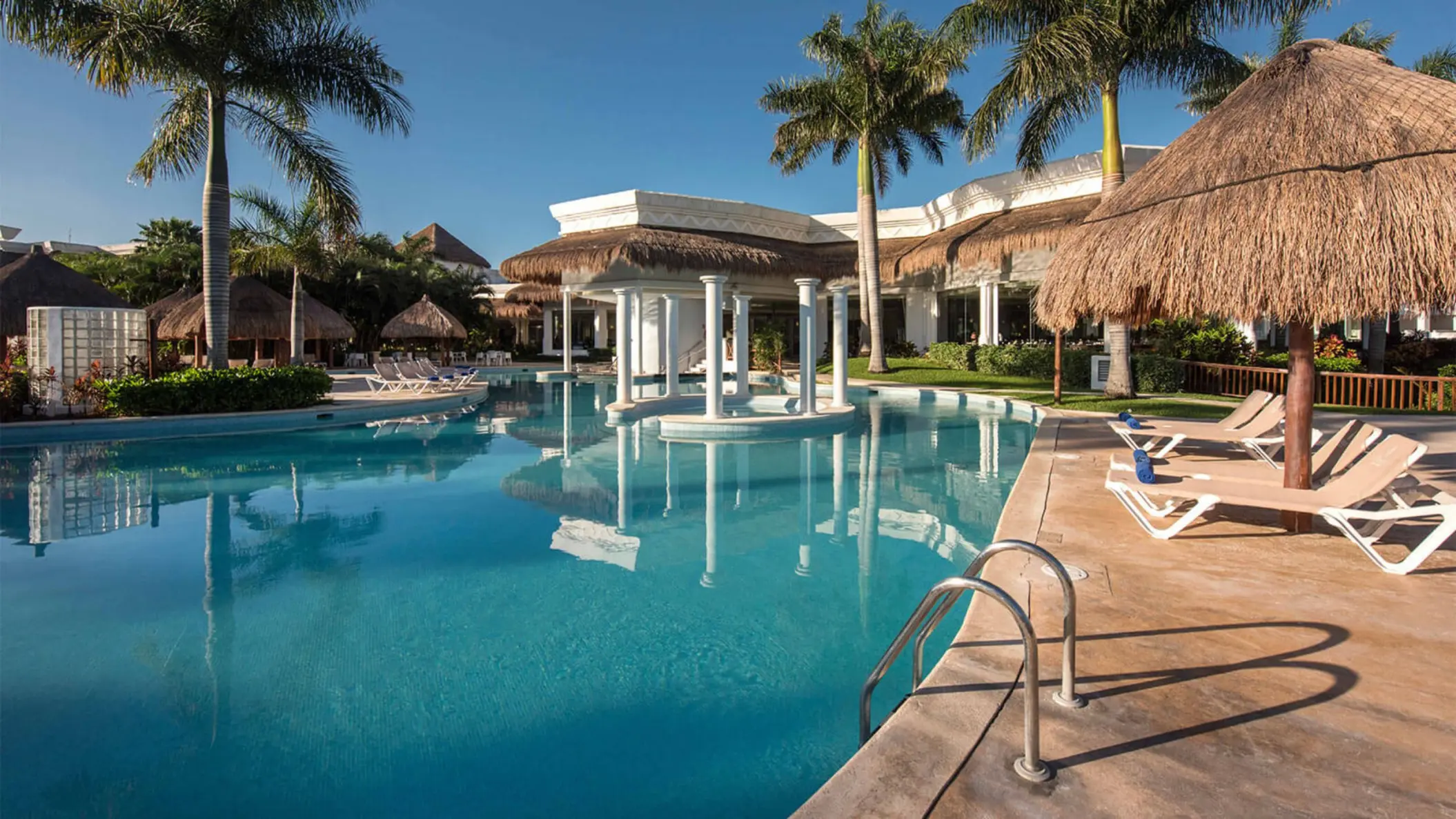 Outdoor swimming pool at Grand Riviera Princess, featuring a central gazebo and numerous lounge chairs under bright blue skies.