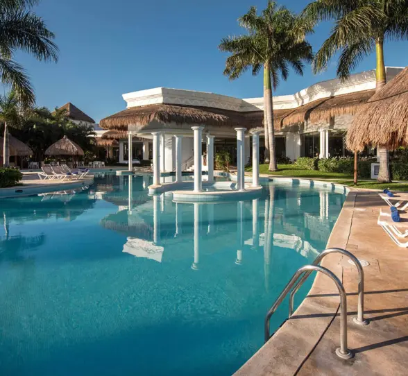 Outdoor swimming pool at Grand Riviera Princess, featuring a central gazebo and numerous lounge chairs under bright blue skies.
