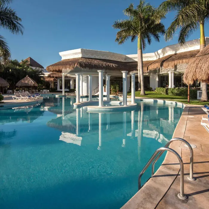 Outdoor swimming pool at Grand Riviera Princess, featuring a central gazebo and numerous lounge chairs under bright blue skies.
