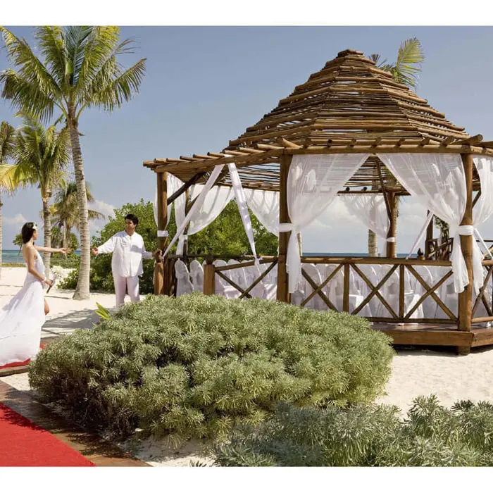 Bride in white gown approaches groom at a beachfront wedding gazebo with white drapes, palm trees, and ocean.