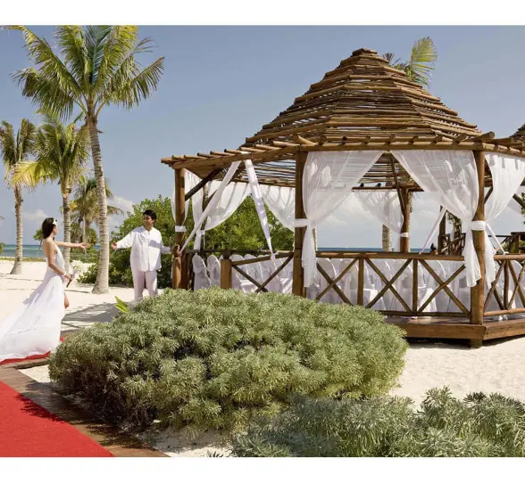 Bride in white gown approaches groom at a beachfront wedding gazebo with white drapes, palm trees, and ocean.