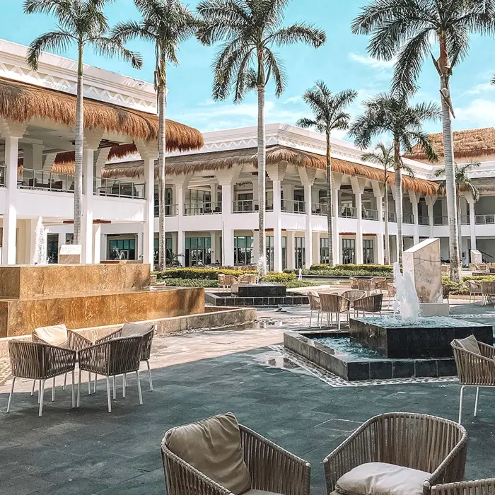 Exterior of Grand Sunset Princess resort with white buildings, palm trees, outdoor seating, and decorative fountains.