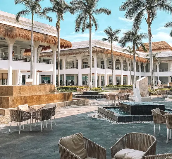 Exterior of Grand Sunset Princess resort with white buildings, palm trees, outdoor seating, and decorative fountains.