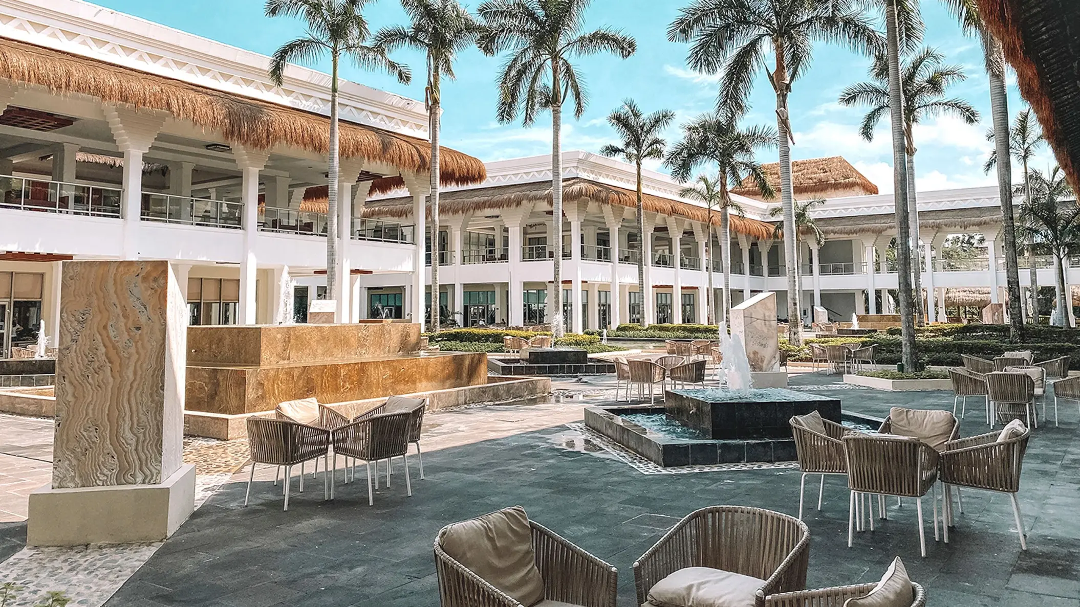 Exterior of Grand Sunset Princess resort with white buildings, palm trees, outdoor seating, and decorative fountains.