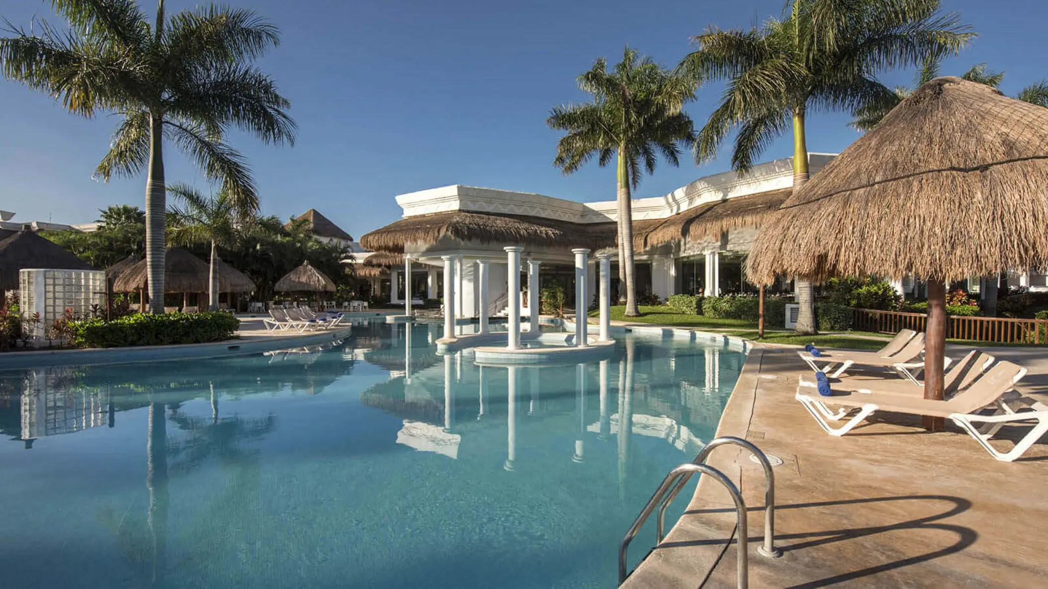 Serene pool at Grand Sunset Princess with a white structure, palm trees, thatched umbrellas, and resort buildings in background.
