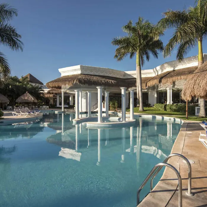 Serene pool at Grand Sunset Princess with a white structure, palm trees, thatched umbrellas, and resort buildings in background.