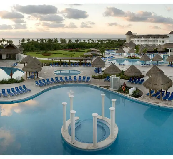 Aerial view of Grand Sunset Princess resort with multiple pools, blue loungers, thatched-roof palapas, and buildings at dusk.