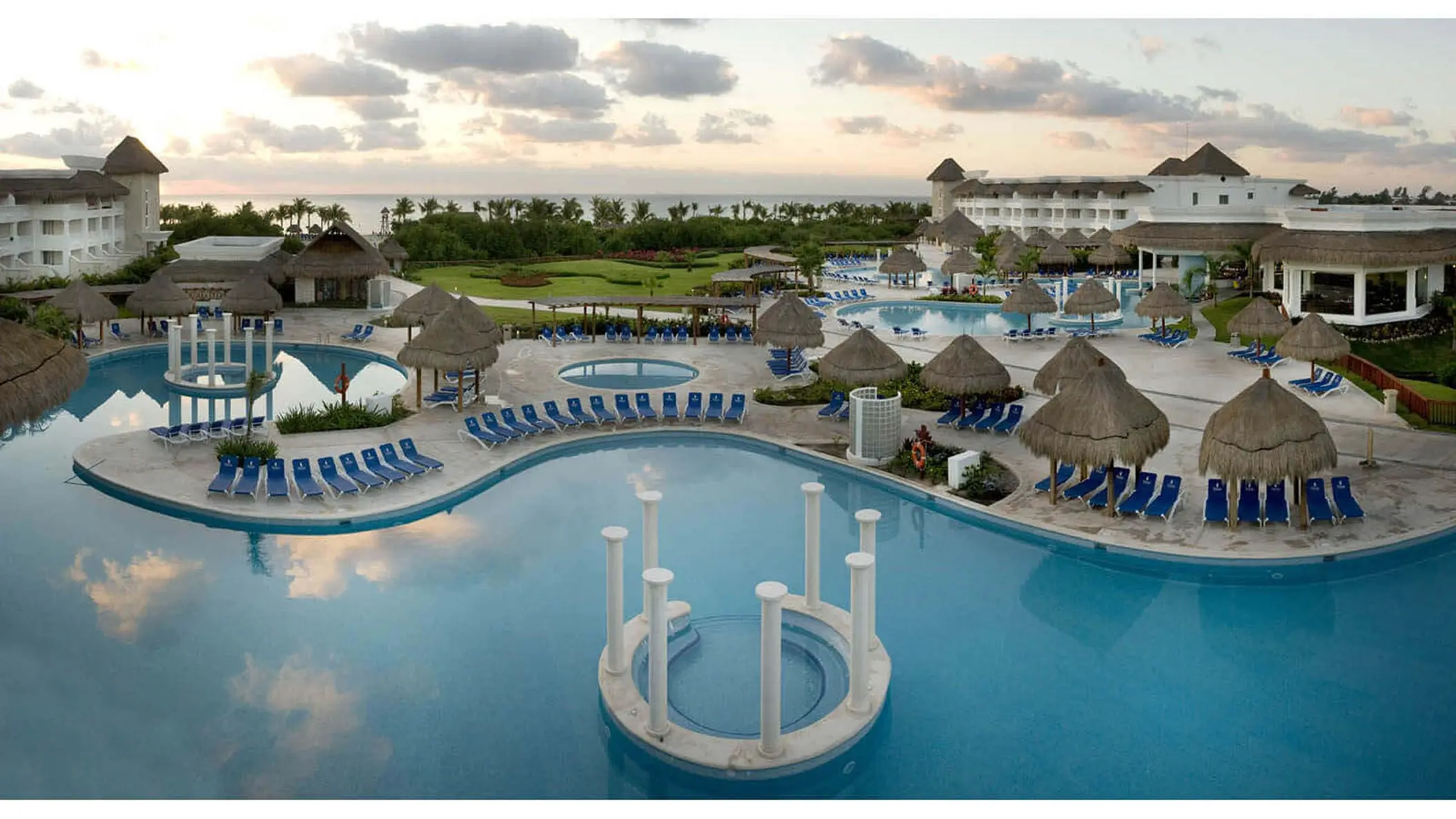 Aerial view of Grand Sunset Princess resort with multiple pools, blue loungers, thatched-roof palapas, and buildings at dusk.