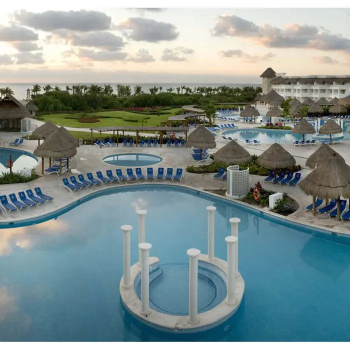 Aerial view of Grand Sunset Princess resort with multiple pools, blue loungers, thatched-roof palapas, and buildings at dusk.