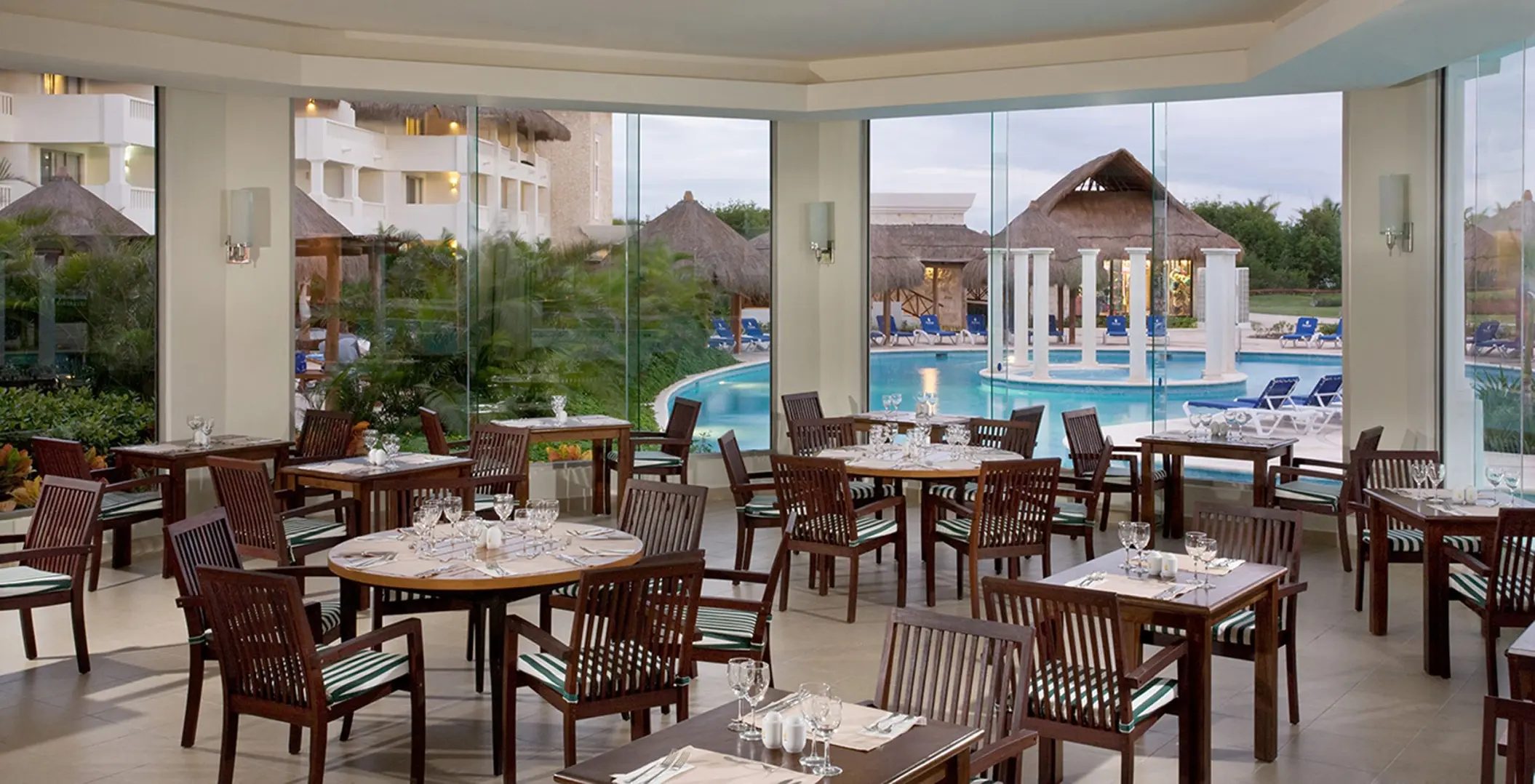 Bright poolside restaurant at Grand Sunset Princess with wooden tables, chairs, and large windows overlooking a pool and palm trees.