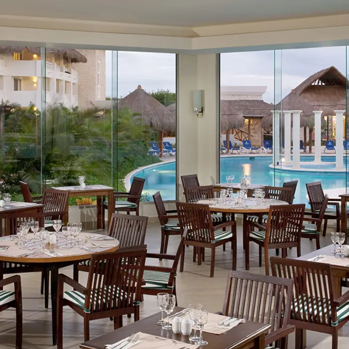Bright poolside restaurant at Grand Sunset Princess with wooden tables, chairs, and large windows overlooking a pool and palm trees.