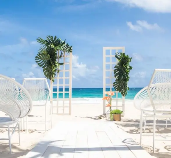 A white-themed wedding ceremony setup on a sandy beach with an arch and chairs facing the ocean.