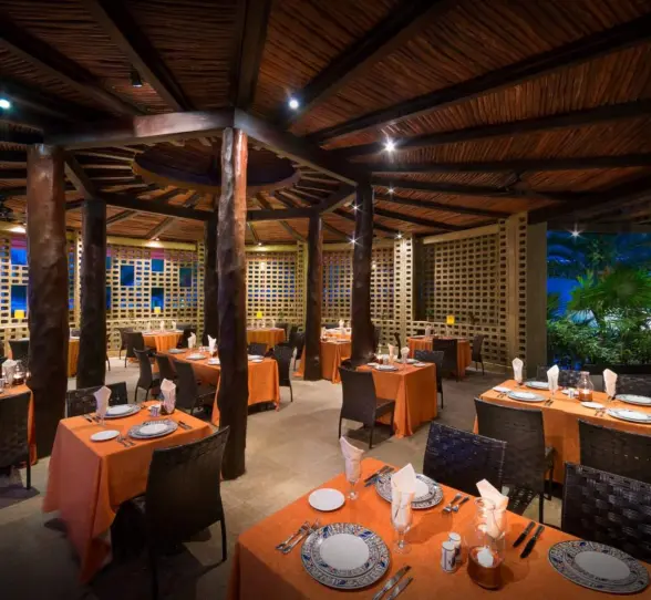 A vibrant, semi-open air restaurant with rustic wood columns and orange-clothed tables set for dinner.
