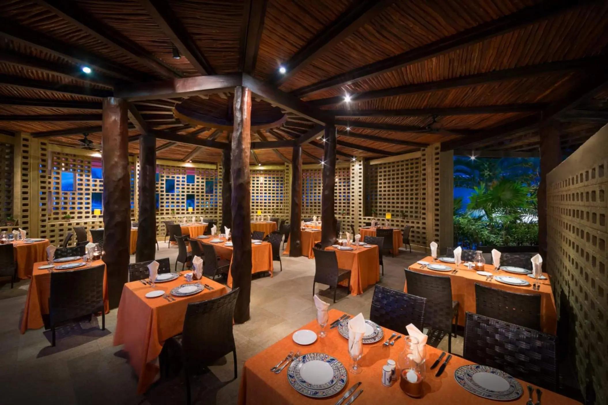 A vibrant, semi-open air restaurant with rustic wood columns and orange-clothed tables set for dinner.