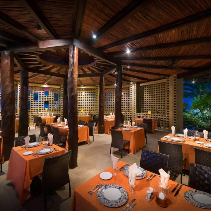 A vibrant, semi-open air restaurant with rustic wood columns and orange-clothed tables set for dinner.