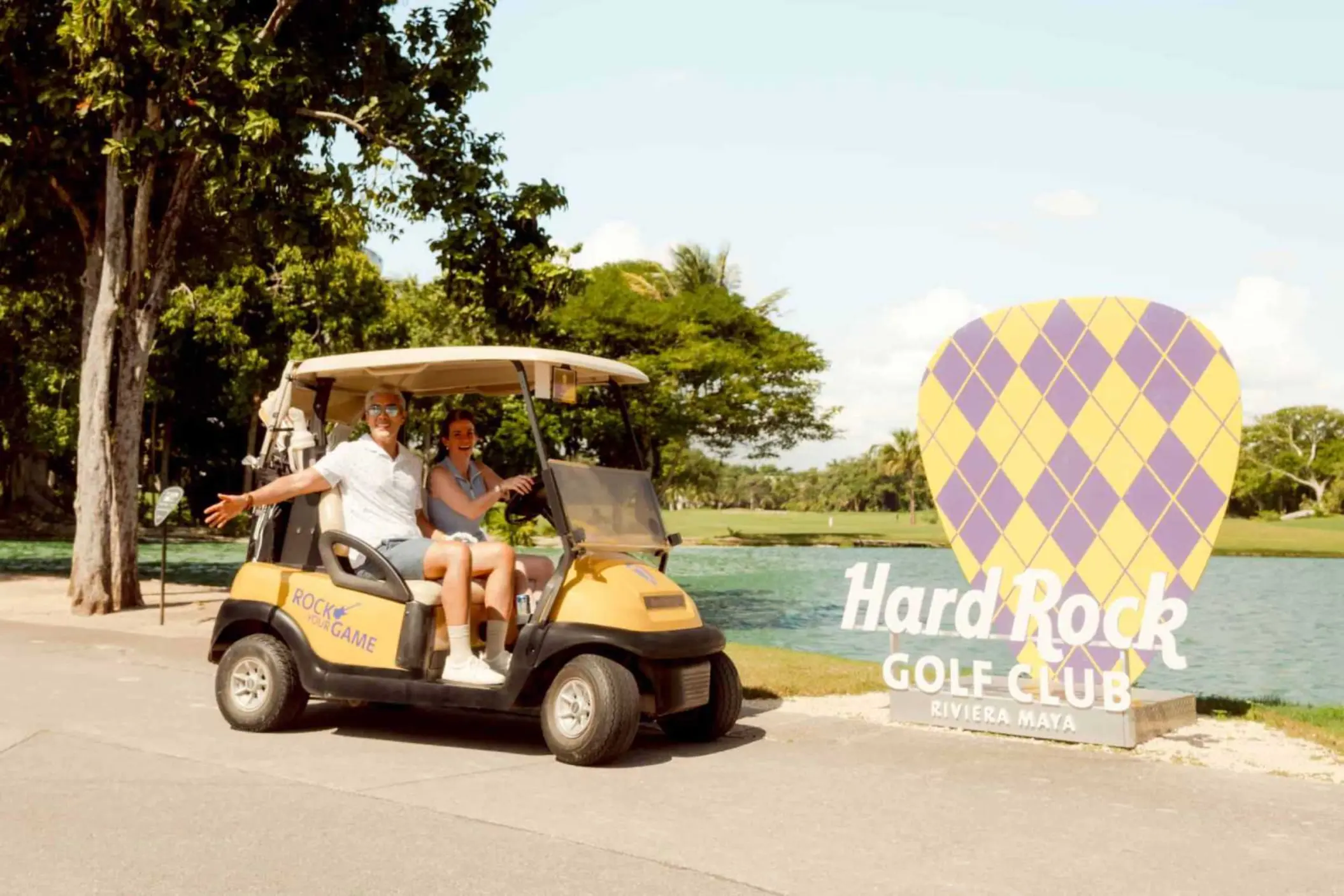 A couple in a golf cart, smiling and waving, next to a sign for the Hard Rock Golf Club.