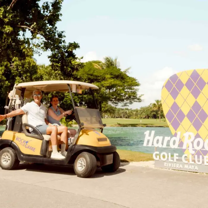A couple in a golf cart, smiling and waving, next to a sign for the Hard Rock Golf Club.