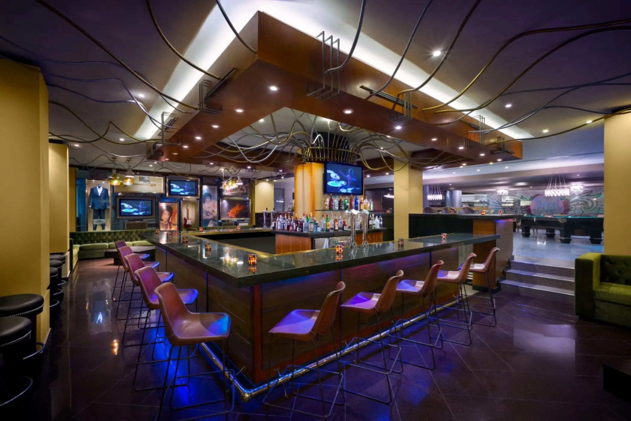 A dimly lit, modern lobby bar with a long counter, multiple TVs, and ambient purple lighting.