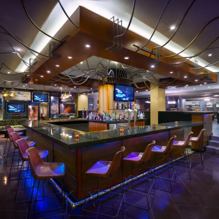 A dimly lit, modern lobby bar with a long counter, multiple TVs, and ambient purple lighting.