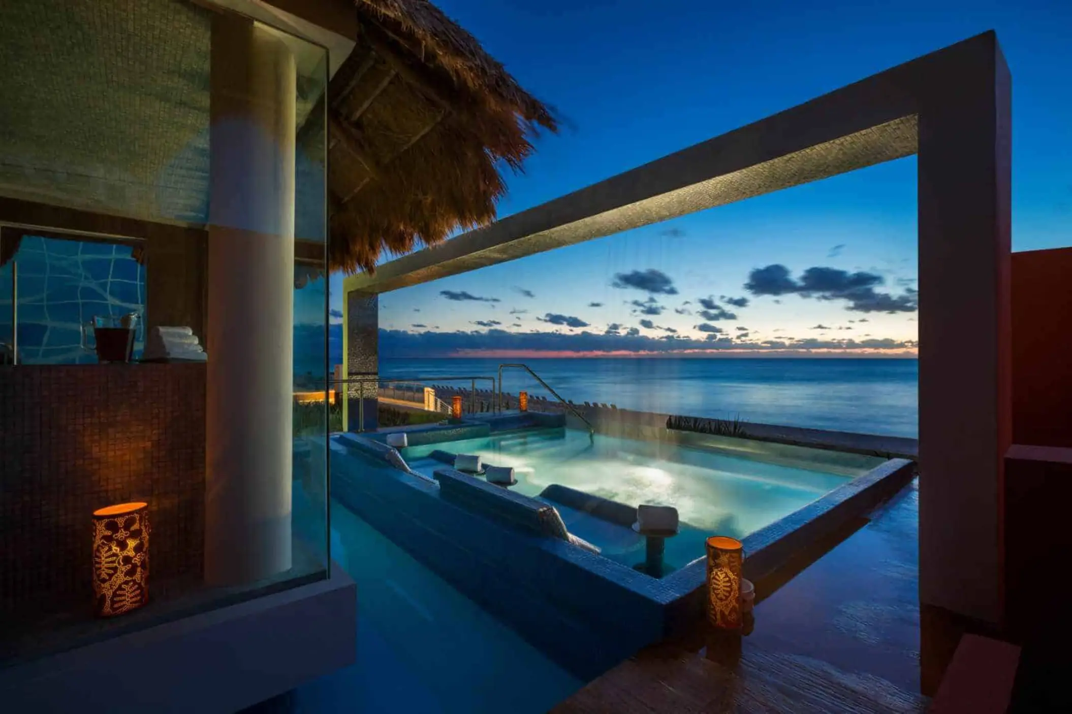 A serene spa hydro-jet pool with a thatched roof, overlooking the ocean at sunset.